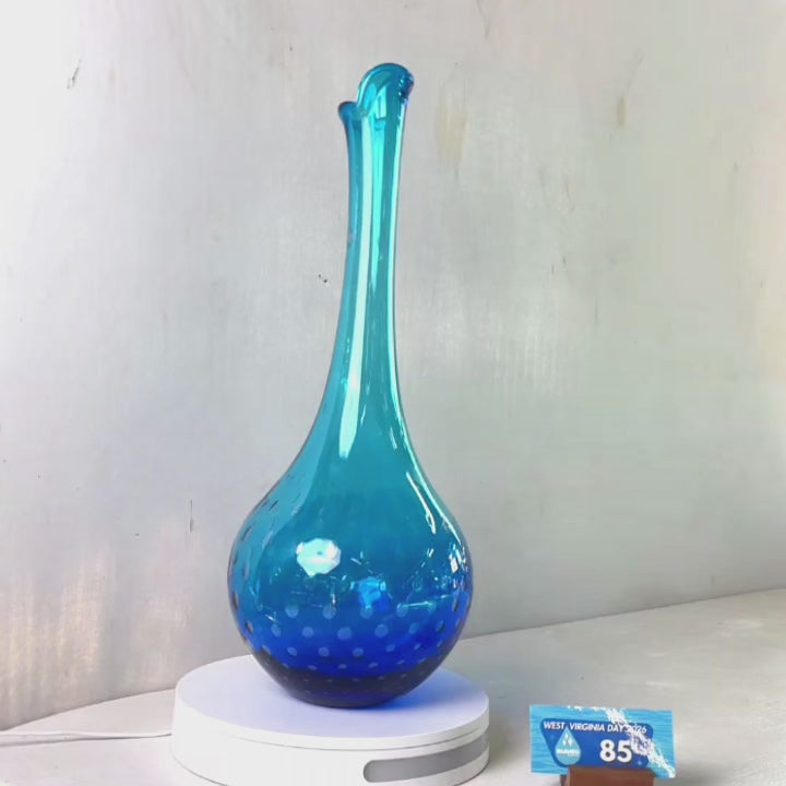 A translucent blue glass vase with a flared rim and subtle white speckled pattern near the base, displayed on a white round scale with a small price tag in front, against a plain light gray wall.
