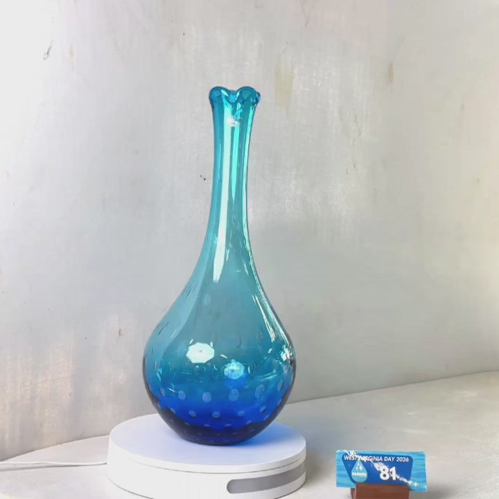 A translucent blue glass vase with a flared rim and subtle white speckled pattern near the base, displayed on a white round scale with a small price tag in front, against a plain light gray wall.