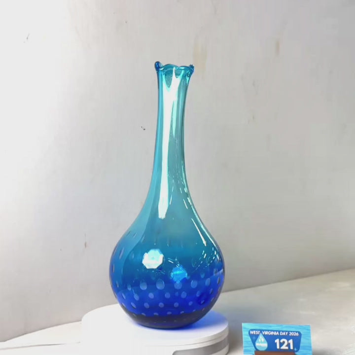 A translucent blue glass vase with a flared neck and dotted pattern near the base, displayed on a white circular light box. A small wrapped item and a brown block are placed beside it on a white surface against a plain light gray wall.