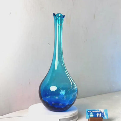 A translucent blue glass vase with a flared rim and dotted pattern near the base, displayed on a white circular stand against a plain light gray wall.