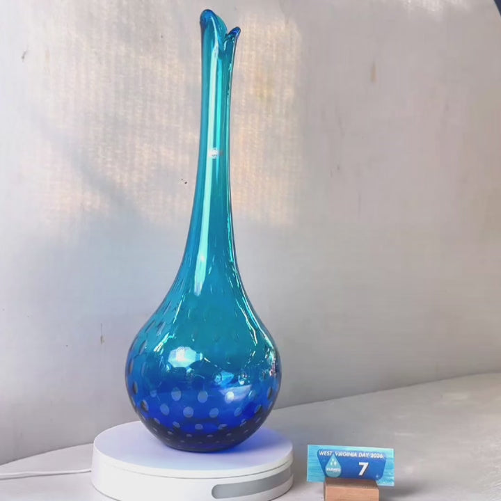 A translucent blue glass vase with a flared neck and dotted pattern, displayed on a white circular base against a light gray wall.
