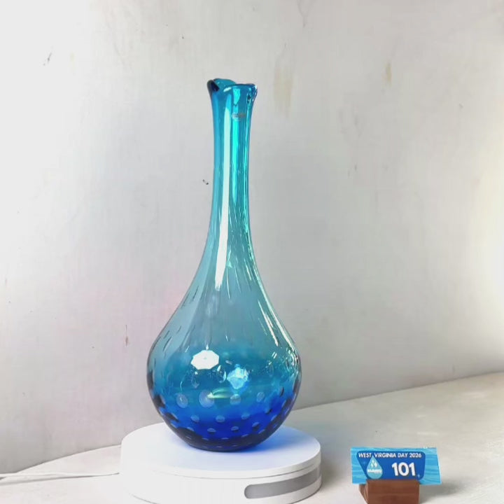 A translucent blue glass vase with a flared rim and dotted pattern near the base, displayed on a white circular stand against a plain light gray wall.