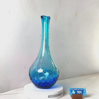 A translucent blue glass vase with a flared rim and dotted pattern near the base, displayed on a white circular stand against a plain light gray wall.