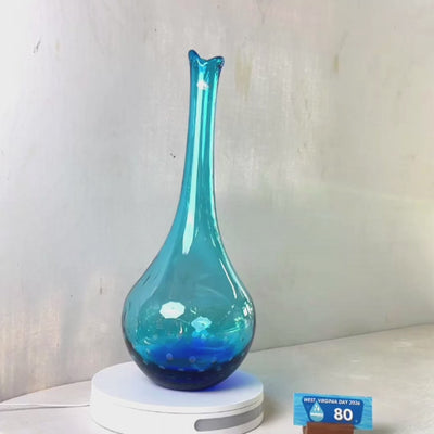 A translucent blue glass vase with a flared rim and subtle white speckled pattern near the base, displayed on a white round scale with a small price tag in front, against a plain light gray wall.