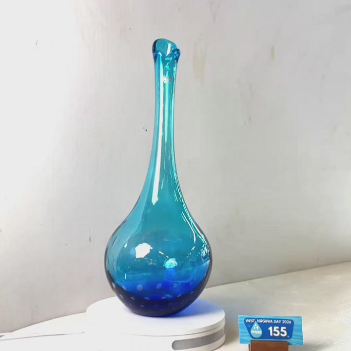 A translucent blue glass vase with a flared rim and dotted pattern near the base, displayed on a white circular stand against a plain light gray wall.