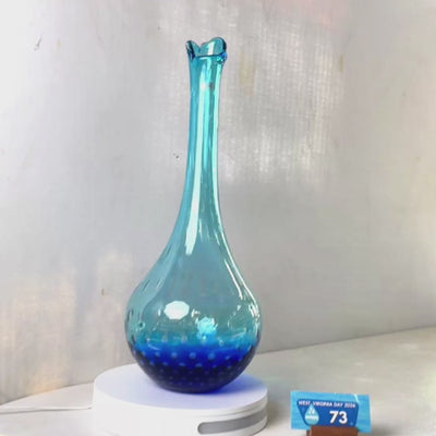 A translucent blue glass vase with a flared rim and subtle white speckled pattern near the base, displayed on a white round scale with a small price tag in front, against a plain light gray wall.