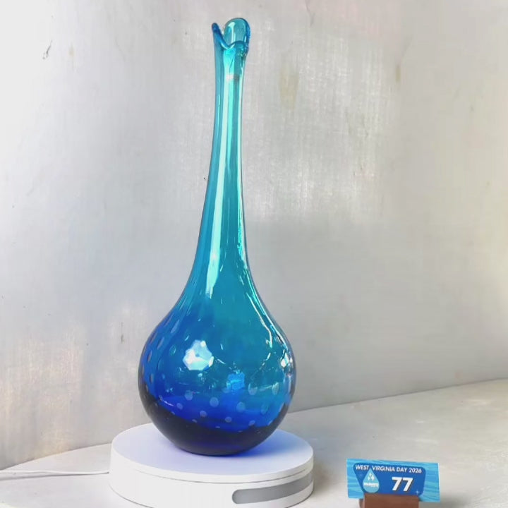 A translucent blue glass vase with a flared rim and subtle white speckled pattern near the base, displayed on a white round scale with a small price tag in front, against a plain light gray wall.