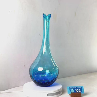 A translucent blue glass vase with a flared rim and dotted pattern near the base, displayed on a white circular stand against a plain light gray wall.