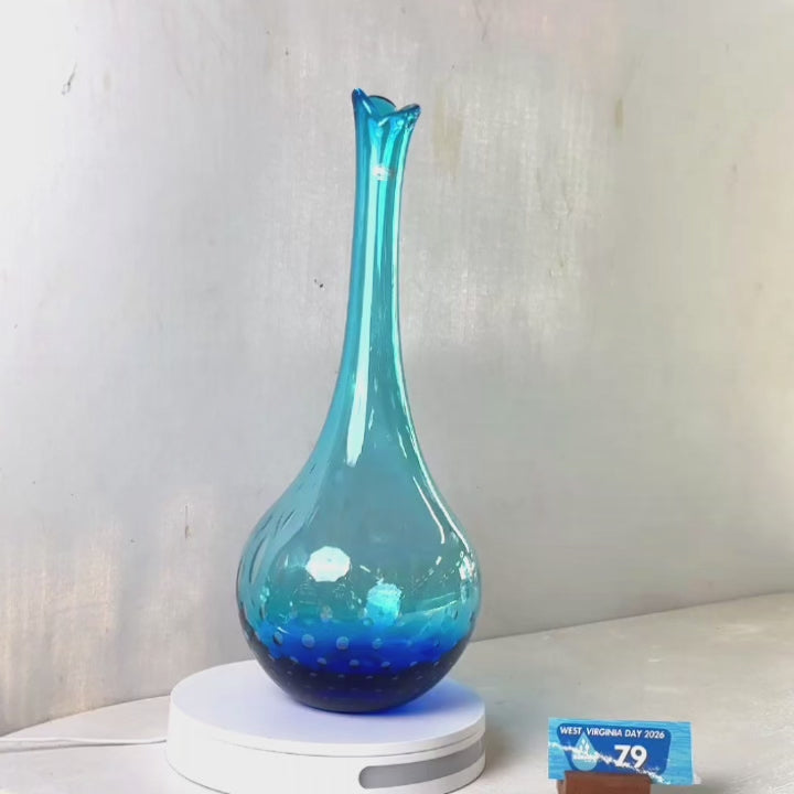 A translucent blue glass vase with a flared rim and subtle white speckled pattern near the base, displayed on a white round scale with a small price tag in front, against a plain light gray wall.