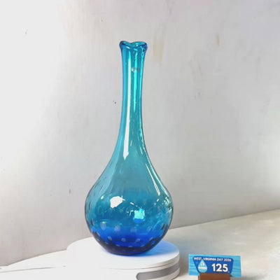 A translucent blue glass vase with a flared neck and dotted pattern near the base, displayed on a white circular light box. A small wrapped item and a brown block are placed beside it on a white surface against a plain light gray wall.