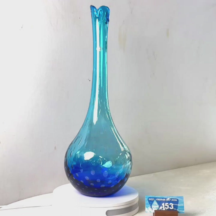 A translucent blue glass vase with a flared rim and dotted pattern near the base, displayed on a white circular stand against a plain light gray wall.