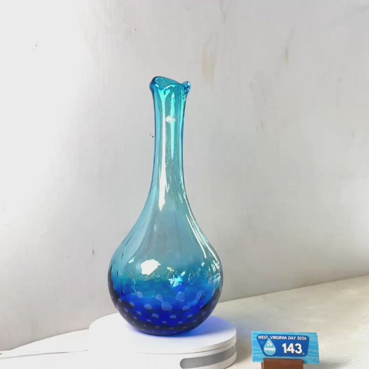 A translucent blue glass vase with a flared rim and dotted pattern near the base, displayed on a white circular stand against a plain light gray wall.