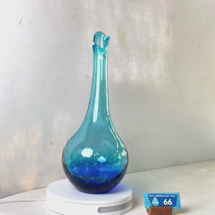 A translucent blue glass vase with a flared neck and dotted pattern, displayed on a white circular base against a light gray wall.