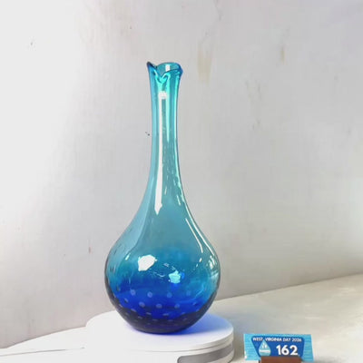 A translucent blue glass vase with a flared rim and dotted pattern near the base, displayed on a white circular stand against a plain light gray wall.