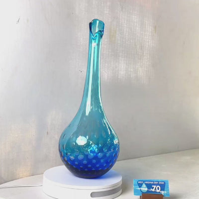 A translucent blue glass vase with a flared neck and dotted pattern, displayed on a white circular base against a light gray wall.