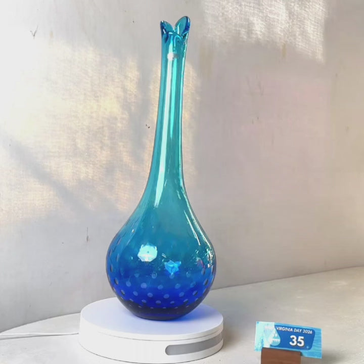 A translucent blue glass vase with a flared neck and dotted pattern, displayed on a white circular base against a light gray wall.