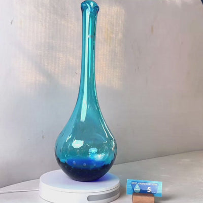 A translucent blue glass vase with a flared neck and dotted pattern, displayed on a white circular base against a light gray wall.