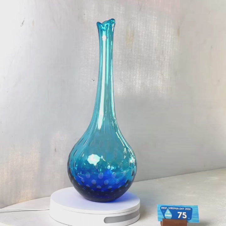 A translucent blue glass vase with a flared rim and subtle white speckled pattern near the base, displayed on a white round scale with a small price tag in front, against a plain light gray wall.