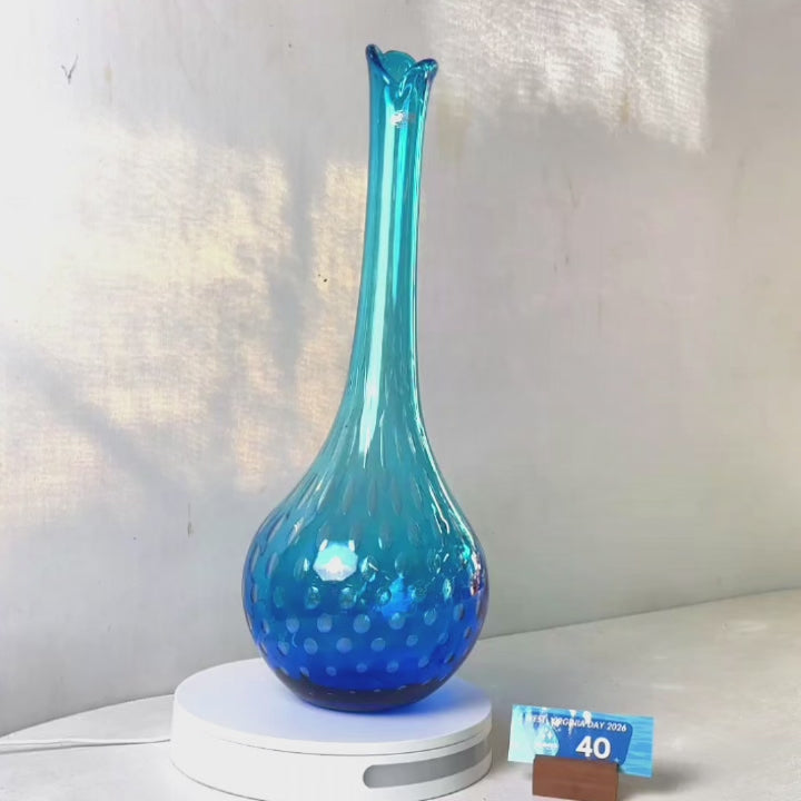 A translucent blue glass vase with a flared neck and dotted pattern, displayed on a white circular base against a light gray wall.